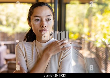 Jeune femme biraciale attentionnée qui regarde loin en se tenant près d'une fenêtre en verre à la maison, l'espace de copie. Style de vie, contemplation et concept de vitiligo. Banque D'Images