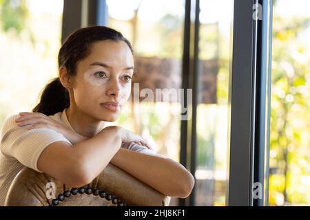 Jeune femme biraciale pensive regardant loin en étant assis sur une chaise à la maison, espace de copie. Style de vie, contemplation et concept de vitiligo. Banque D'Images