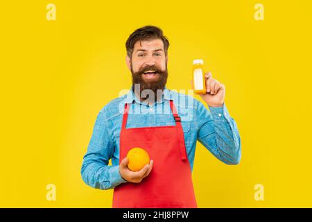 Homme heureux en tablier tenant orange et bouteille de jus fond jaune, barman Banque D'Images