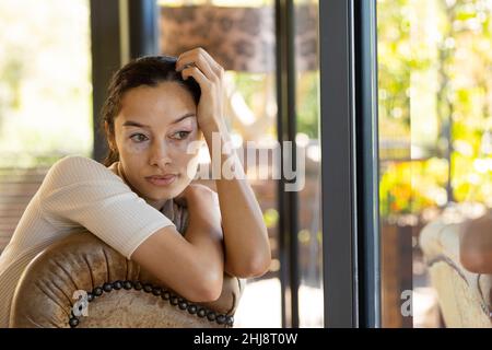 Jeune femme biraciale contemplative avec vitiligo regardant loin tout en étant assis sur une chaise à la maison Banque D'Images