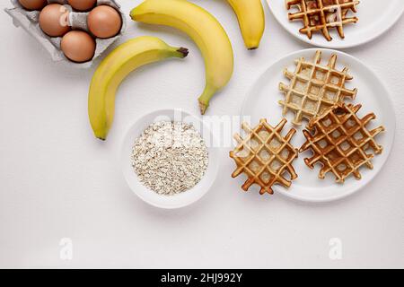 Gaufres maison sans gluten avec banane et flocons d'avoine, ingrédients pour leur préparation sur une table blanche, repas sains et petit déjeuner sans sucre, Banque D'Images