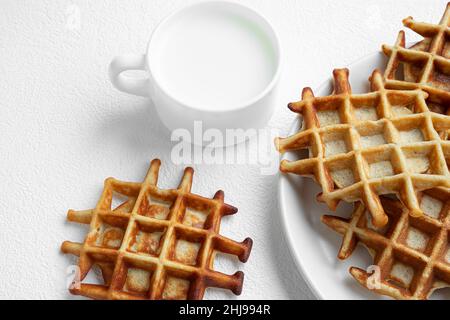 Gaufres belges fraîchement cuites sur une plaque blanche en gros plan et une tasse de lait sur une table blanche, concept de petit déjeuner savoureux Banque D'Images