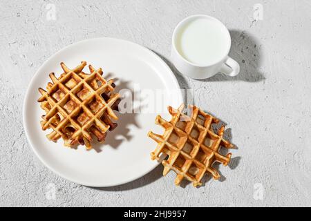Gaufres belges fraîchement cuites sur une assiette blanche et une tasse de lait sur une table légère, concept de petit déjeuner savoureux, vue du dessus Banque D'Images