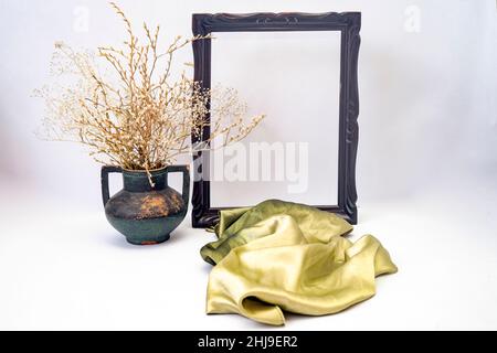 maquette à cadre vide avec foulard vert soyeux et vase vintage avec fleurs et branches séchées Banque D'Images