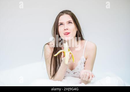 La fille est assise sur le lit, enveloppée dans une couverture, et appréciant manger une banane.Le brunette prend le petit déjeuner avec des fruits. Banque D'Images