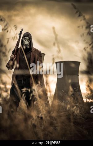 Photo d'un survivant apocalyptique portant un masque à gaz et tenant une arme à feu en se tenant devant une centrale nucléaire dystopique et polluée. Banque D'Images