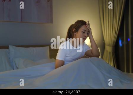 Femme asiatique couchée sur un lit blanc dans la chambre à coucher qui a l'air inquiète ou qui pense à quelque chose sur sa vie ou son travail en milieu de nuit à la maison. Banque D'Images