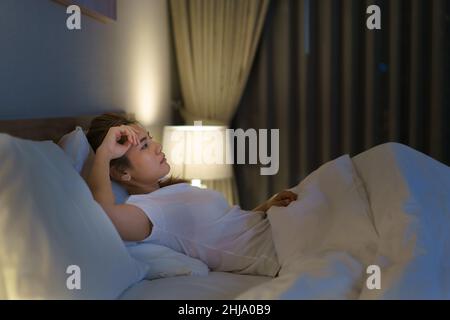 Femme asiatique couchée sur un lit blanc dans la chambre à coucher qui a l'air inquiète ou qui pense à quelque chose sur sa vie ou son travail en milieu de nuit à la maison. Banque D'Images