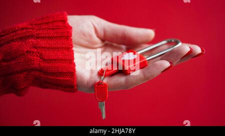 Gros plan de la main d'une femme dans un chandail rouge tenant un cadenas avec un coeur avec des clés sur un fond rouge.le concept de la Saint-Valentin, mariage, un symb Banque D'Images