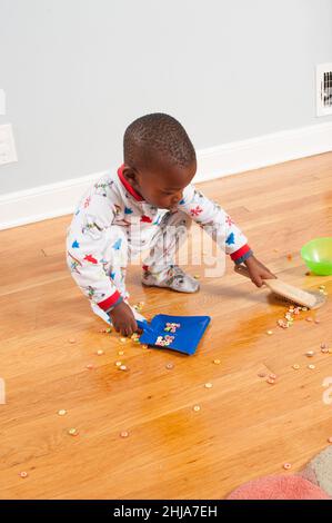 Un tout-petit garçon à la maison utilise un bac à poussière et une brosse pour nettoyer les céréales déversées sur le sol, portant un pyjama Banque D'Images