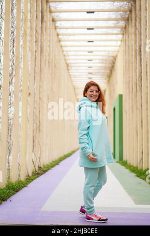 Femme caucasienne aux cheveux rouges dans un pull à capuche à la menthe marchant dans le parc. Banque D'Images