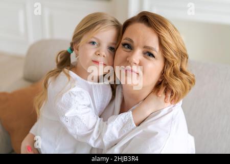 Une mère européenne et une petite fille s'embrasent tout en étant assis sur un canapé dans le salon lumineux de la maison.La relation entre les enfants et les parents.Bonne maternité Banque D'Images