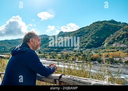 Vue latérale d'un heureux homme mûr et réfléchi contemplant la nature montagneuse avec un ciel nuageux près d'une passerelle en bois.Concept des activités de loisirs Banque D'Images