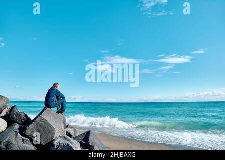 Vue latérale de l'homme mûr barbu assis au sommet de la roche volcanique contemplant la mer bleue.Concept d'activités de loisirs, tourisme, style de vie e Banque D'Images