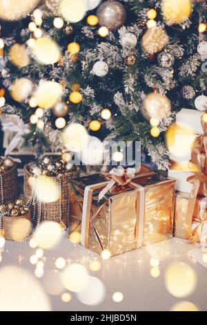 Gros plan de boîte cadeau dorée emballée avec noeud sur le sol sous l'arbre de Noël entouré d'une guirlande d'ampoules jaunes, de nombreux cadeaux de vacances et de Noël arbre de Banque D'Images