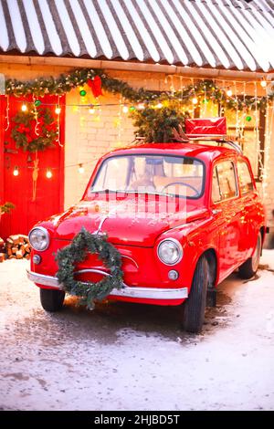 L'esprit des fêtes.Confortable photo de style de Noël avec voiture rétro rouge dans la cour couverte de neige de la maison décorée avec des branches de pin et des lumières de Noël.Sortie Banque D'Images