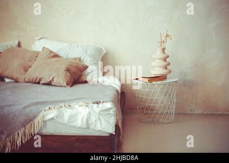 Décoration élégante et minimaliste de style loft avec murs en béton clair, très grand lit en bois avec oreillers, couverture et table de chevet ronde en métal Banque D'Images