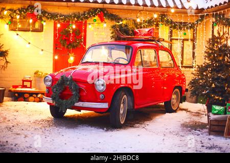 L'esprit des fêtes.Confortable photo de style de Noël avec voiture rétro rouge dans la cour couverte de neige de la maison décorée avec des branches de pin et des lumières de Noël.Sortie Banque D'Images