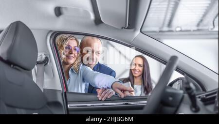 Épouse montre à son mari les caractéristiques d'une nouvelle voiture dans la concession, souriant, tandis que l'assistant commercial se tient de côté. Banque D'Images