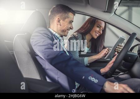 Un homme qui teste une voiture dans le magasin sourit en regardant une tablette dans les mains de la femme concessionnaire de voiture montrant des aspects techniques. Banque D'Images