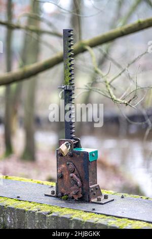 Pièce en métal vieille et rouillée avec un engrenage à engrenages dans une porte de dérivation sur un pont, fixé avec un cadenas pour qu'il ne soit pas utilisé, des branches d'arbres et un ruisseau dans un mi Banque D'Images