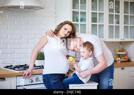 une famille de trois personnes avec une femme enceinte et un jeune fils s'embrasse dans une grande cuisine blanche Banque D'Images