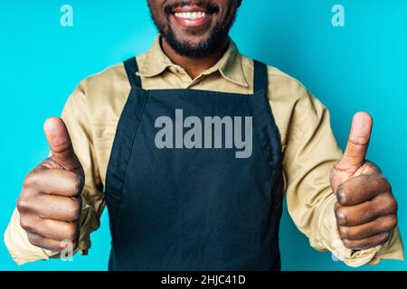 joyeux latin homme en coton chemise ant tablier noir dans le studio bleu regardant l'appareil photo et se sentir bien Banque D'Images