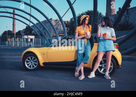 Les jeunes femmes sont souriantes, appréciant les frites provenant de l'emballage en papier et les boissons dans des gobelets en papier, se posant près de la voiture jaune.Restauration rapide.Copier l'espace Banque D'Images