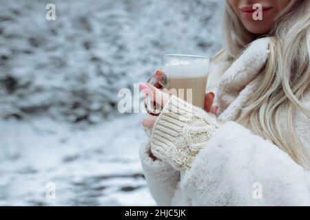 De belles mains tiennent une tasse de cappuccino sur le fond de la forêt d'hiver.Gants doux beige sur les mains d'une fille dans les mains de Banque D'Images