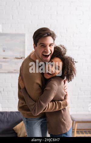 Un couple multiethnique positif s'embrasse et regarde la caméra à la maison Banque D'Images