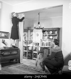 Dans le 1940s.Un jeune couple organise les rideaux qui divisent les chambres de leur appartement.Mme Vibeke Falk se tient sur un meuble pour atteindre les clips et ajuster les draperies tandis que M. Lauritz Falk et leur fille regarde.La chambre à l'arrière possède un intérieur rustique nordique avec un mobilier traditionnel en bois et un arrière-plan historique.Suède 1946 Kristoffersson réf. Y38-4 Banque D'Images