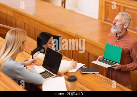 Un enseignant mature tenant un ordinateur portable avec écran vert près des étudiants interraciaux de l'université Banque D'Images