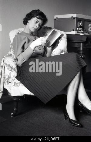 1950s, historique, à l'intérieur d'une pièce, assis dans une chaise, une jeune femme élégamment habillée portant un chemisier en laine et une jupe plissée, lisant un livre de poche sur le peintre italien Botticelli, avec un lecteur de disques portable sur une table à côté d'elle, Angleterre, Royaume-Uni Banque D'Images