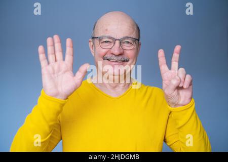 Homme senior levant la main, montrant le numéro sept avec les doigts, comptant quelque chose, debout sur fond bleu Banque D'Images