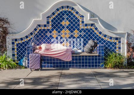 Une femme sans abri dort avec une couverture rose à midi sur un banc recouvert de tuiles fantaisie sur un trottoir à Santa Barbara, Californie. Banque D'Images