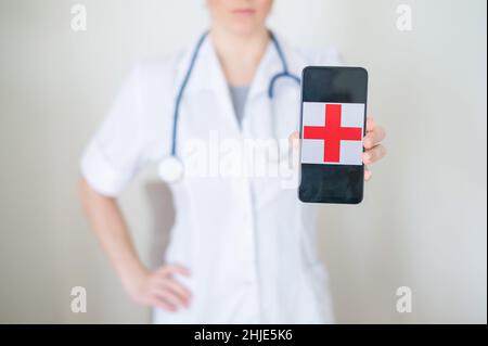 Une femme médecin sans visage tient un smartphone avec une croix rouge sur l'écran. Infirmière méconnaissable en manteau médical faisant la démonstration d'un téléphone cellulaire. Banque D'Images