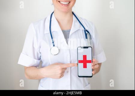 Une femme médecin méconnaissable tient un smartphone avec une croix rouge sur l'écran.L'infirmière sourit et pointe vers l'écran d'un téléphone cellulaire. Banque D'Images