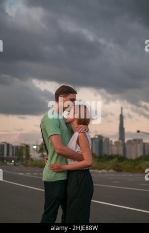 Charmant jeune couple debout dans la rue de la métropole sur le coucher du soleil et de l'eau.Petit ami embrasse petite amie.Gratte-ciel en arrière-plan Banque D'Images