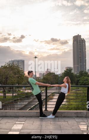 Charmant couple caucasien debout sur le pont au coucher du soleil, tenant les mains et regardant l'un l'autre.Petite amie et petit ami. Banque D'Images