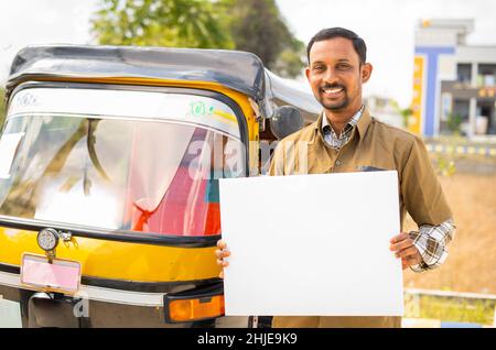 Un chauffeur d'auto souriant avec un panneau blanc vide regardant la caméra en se tenant à côté de la caméra - convecteur de publicité et de bien-être Banque D'Images