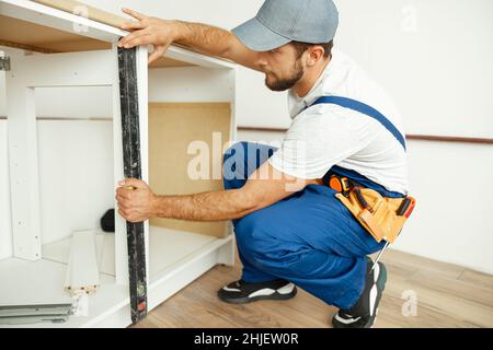 Homme de main professionnel, homme ouvrier en uniforme en utilisant le niveau à bulle tout en vérifiant les mesures de l'armoire de cuisine à l'intérieur Banque D'Images