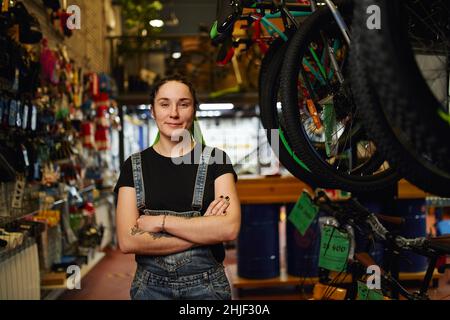 Contenu informel jeune femme vendeur en denim général avec tatouages sur les bras et les dreadlocks souriant et regardant l'appareil photo tout en se tenant près de la bik moderne Banque D'Images