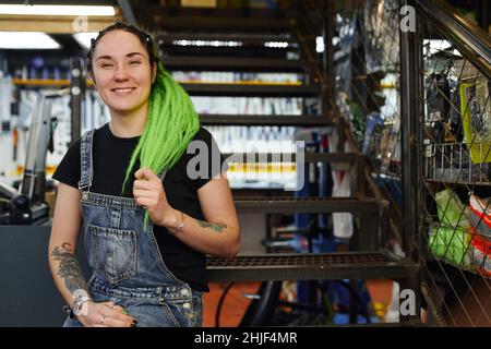 Femme optimiste vendeur avec des dreadlocks lumineux assis sur l'escalier dans le magasin vendant des marchandises et des outils de vélo et souriant à la caméra Banque D'Images
