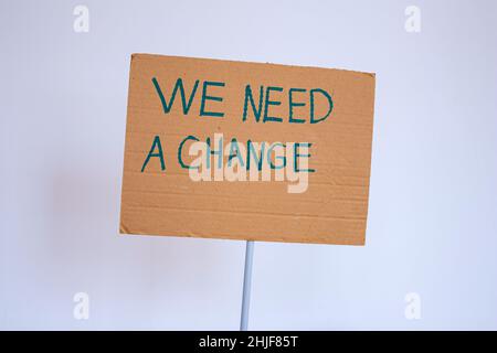 Main tenant le carton avec l'inscription « nous avons besoin d'un changement » sur fond gris de mur.Elle proteste peut-être contre le réchauffement climatique ou la pollution de l'environnement. Banque D'Images