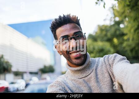 Homme afro-américain en gilet à col roulé et lunettes prenant un selfie dans la ville Banque D'Images