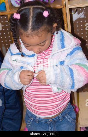 Classe préscolaire 4-5 ans auto-soin fille zipping propre veste, reliant les deux parties de la fermeture éclair, debout devant le cubby Banque D'Images