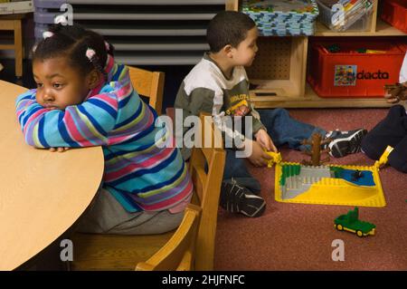 Éducation classe préscolaire 4-5 ans triste, dissipe fille assise seule à la table comme d'autres enfants jouent en arrière-plan Banque D'Images