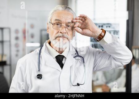 Un médecin vieillissant et attentif en blouse de laboratoire et en lunettes regardant de côté pendant qu'il se tient au bureau de l'hôpital.Portrait d'un travailleur médical qualifié. Banque D'Images