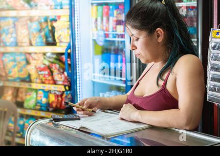 latina femme se concentrant tout en calculant le nombre de ventes dans un carnet de son magasin d'épicerie. fille penchée sur une vitrine de verre pensant abo Banque D'Images