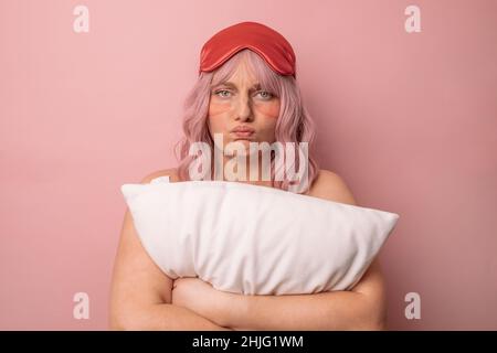 Contrarié endormi la femme caucasienne porte un masque de sommeil, triste de se réveiller tôt le matin pose isolée sur fond rose. Banque D'Images